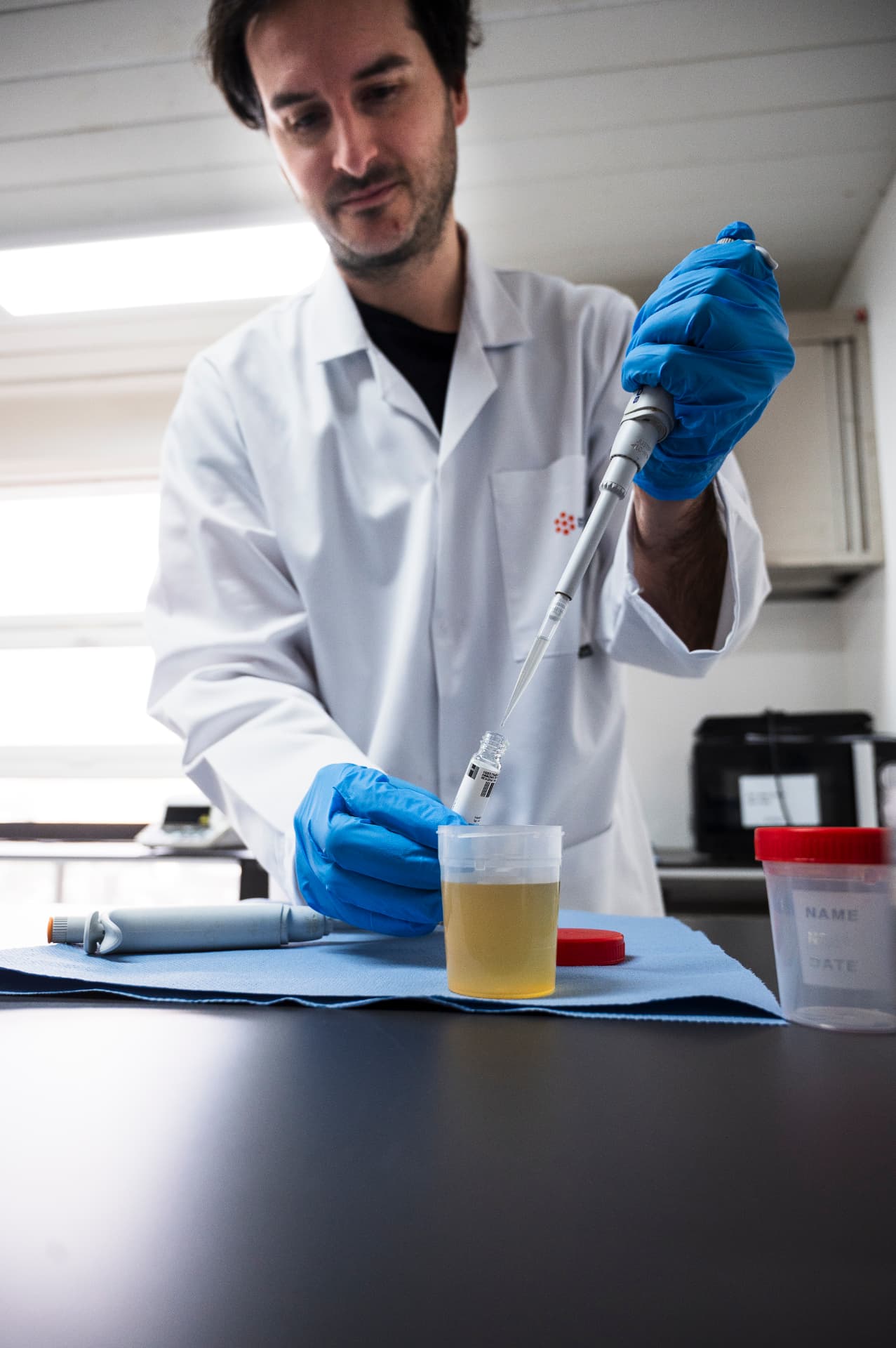 Medewerker in een labojas met blauwe handschoenen pipetteert een geel staal in een reageerbuis