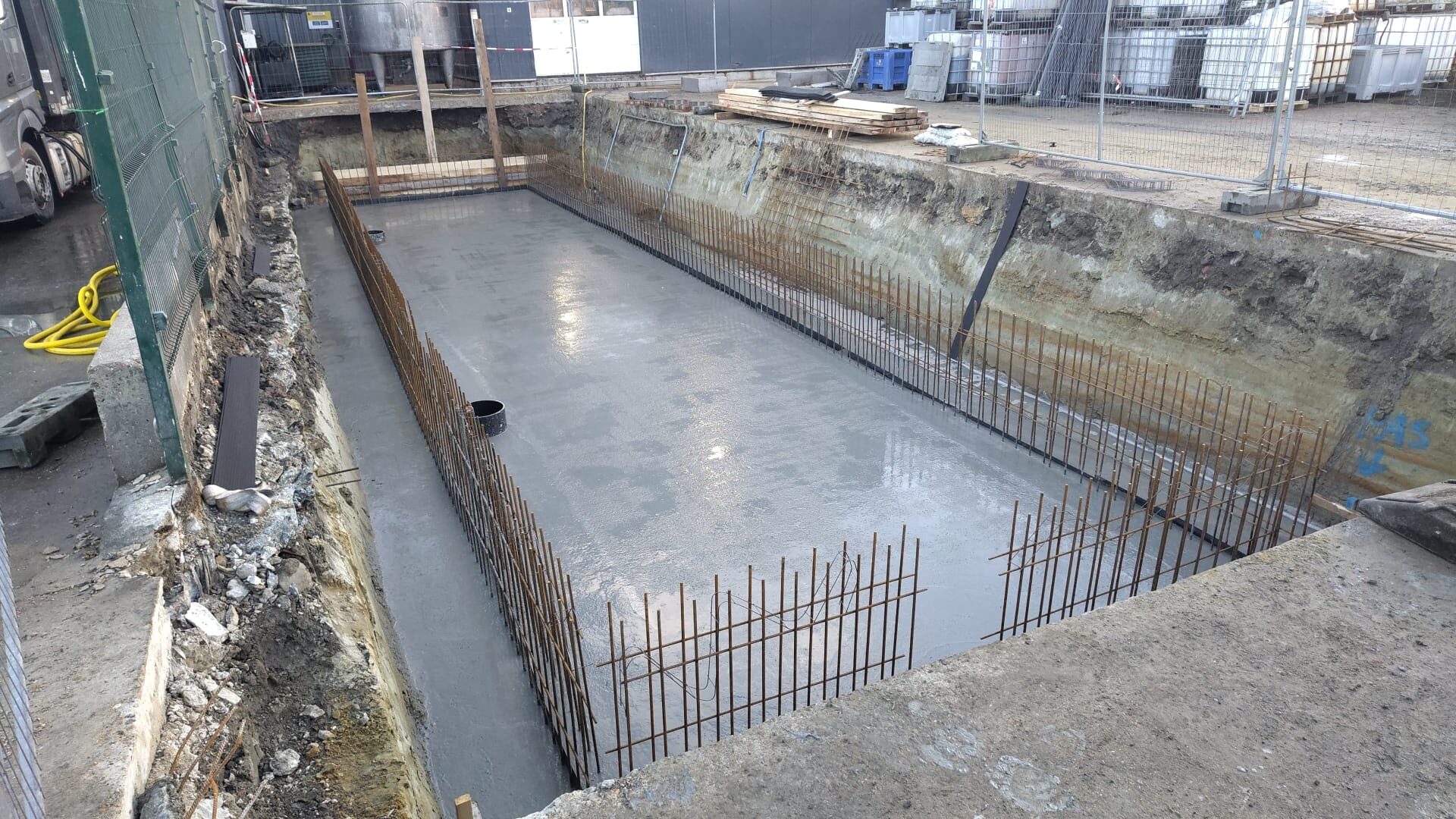 Een uitgegraven bouwput met een vers gestorte betonvloer en rechtopstaande wapeningsstaven langs de randen.