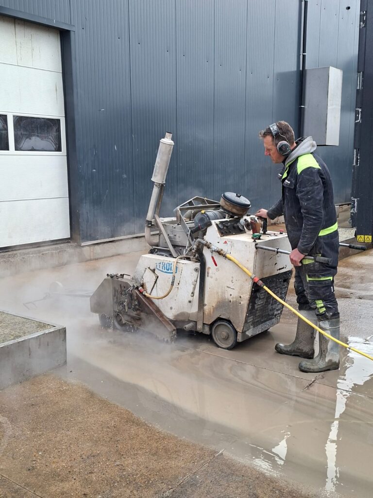 Een arbeider met gehoorbescherming zaagt met een betonzaagmachine in de natte betonvloer naast een loods.