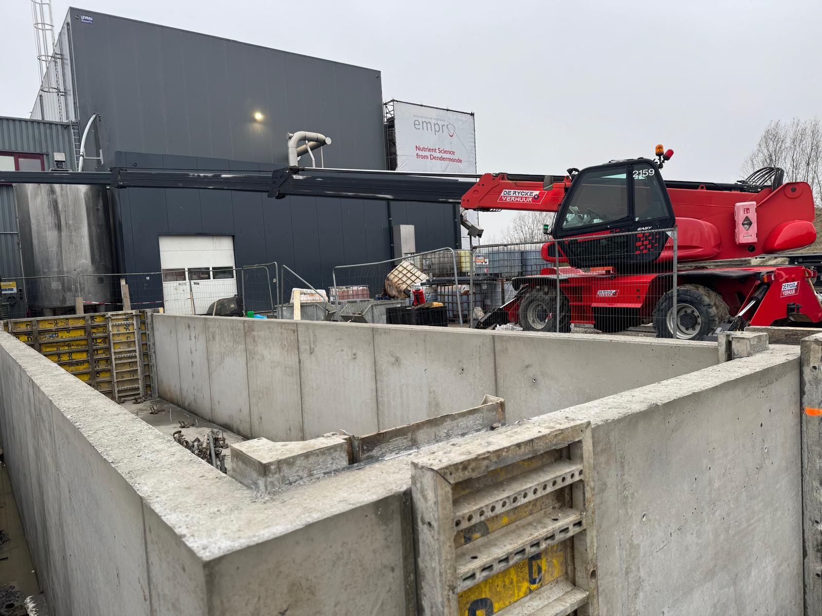 Nieuwe betonnen muren van de bouwput, met op de achtergrond een rode verreiker en het bedrijfsgebouw met het Empro-logo.